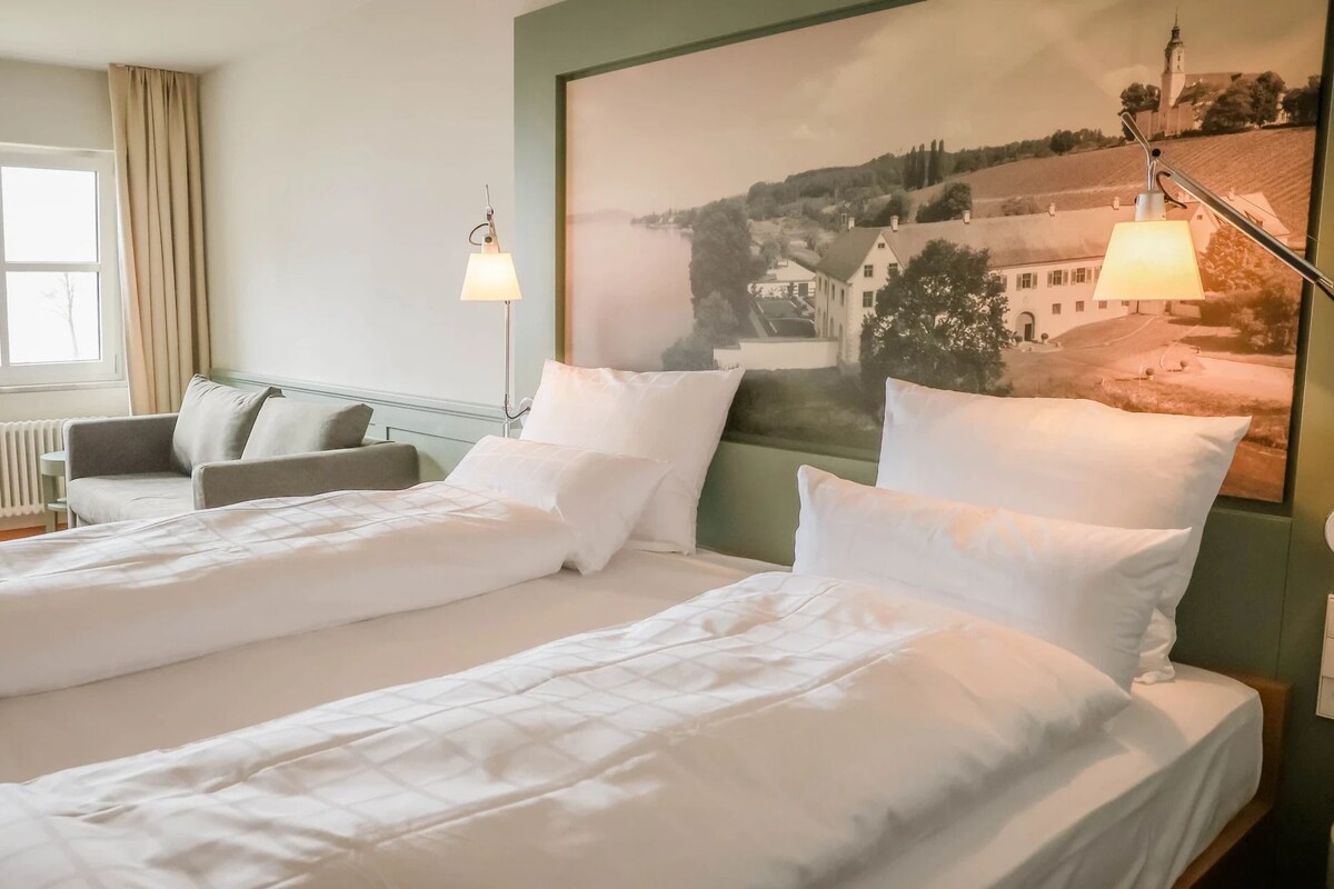 A hotel room with two beds, a sofa, a radiator, a window with curtains, and a large landscape painting on the wall.