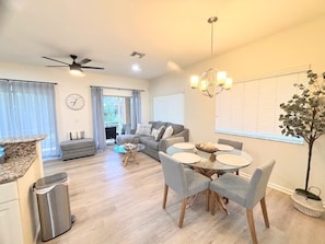 Un salon moderne comprenant un canapé, une table à manger et des chaises, un ventilateur de plafond et une grande fenêtre avec des stores.