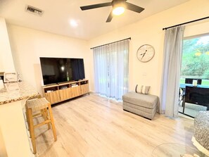 Ein Wohnzimmer mit Flachbildfernseher, einer Couch, einem hölzernen Hocker, einem Deckenventilator und einem Fenster mit Vorhängen.