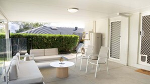 A covered outdoor area with a sofa, chairs, a round table, and an air conditioning unit. - 4BR Luxury Retreat (Gold Coast)