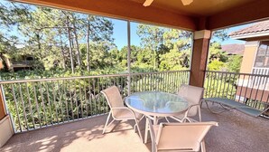 Un patio cubierto con una mesa y sillas de vidrio, baranda de metal y vista a árboles y un edificio.