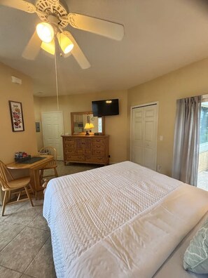 Ein Schlafzimmer mit Bett, Schreibtisch, Stuhl, Kommode, Deckenventilator, Fernseher und einem Fenster mit Vorhängen.