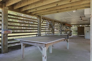 Uma mesa de pingue-pongue sob uma estrutura de madeira com telhado, rodeada por ripas de madeira e um piso de concreto.