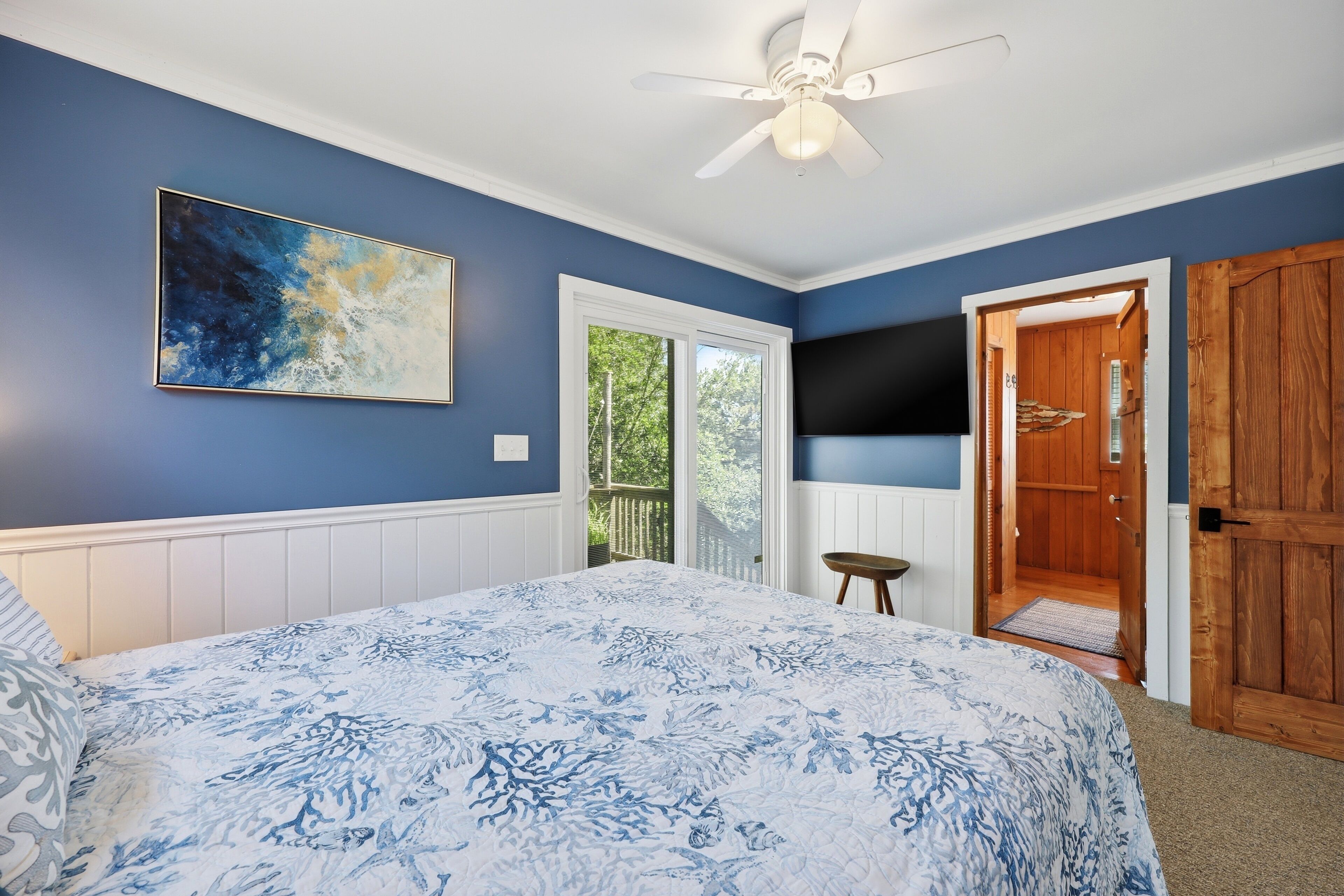 Ein Schlafzimmer mit einem Bett, einem Deckenventilator, einem Gemälde, einem Fernseher und einer Holztür.