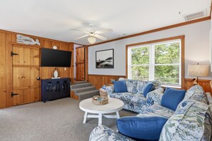 A living room with a wooden wall, a ceiling fan, a flat-screen TV, a sofa, a coffee table, and a window with blinds. - 3BR Duck cottage in Sound Sea, near the beach and Duck village. (Duck)
