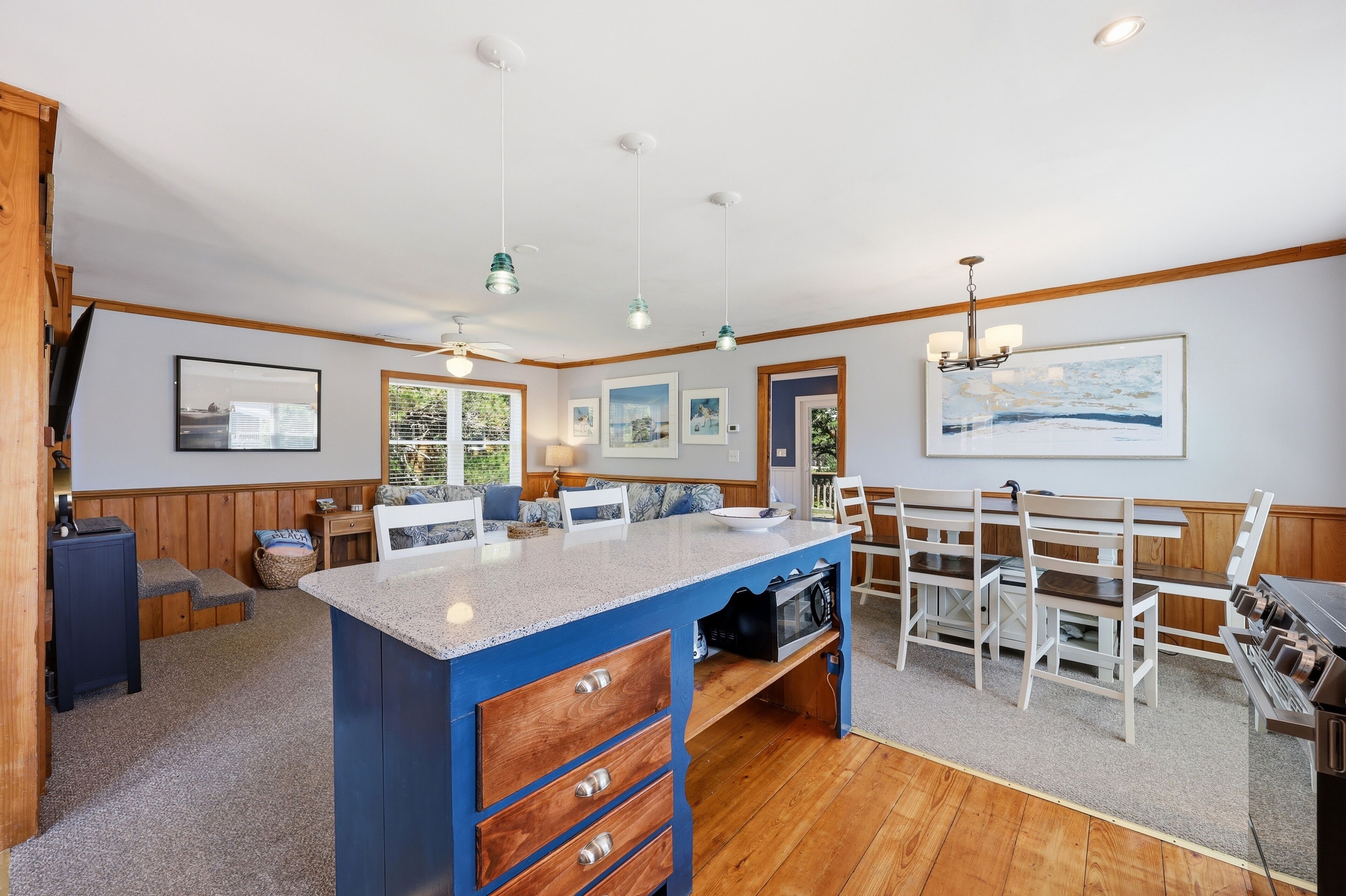 A kitchen with a blue island, wooden cabinets, a dining area with white chairs, and a large window.