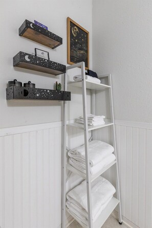 A white metal shelf unit with folded white towels and decorative items on shelves. - Cowboys and Fairytales Movie Theater  Games (Abilene)