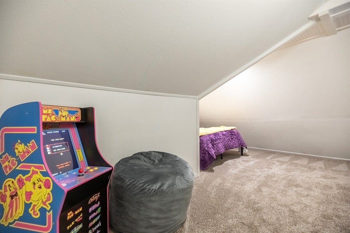 A room with a single arcade game, a bean bag chair, and a bed with a purple blanket.
