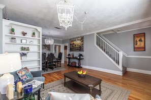 Ein Wohnzimmer mit Sofa, Couchtisch und einer Chandelüster. Im Hintergrund ist eine Treppe und ein Essbereich zu sehen.