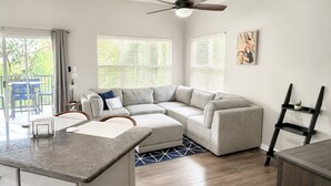 A modern living room with a grey sectional sofa, a dining area with a table and chairs, and a balcony with blue chairs. - Right Direction Rentals 1 Bedroom Villa 9887 (Port St. Lucie)