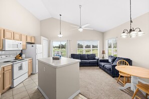 A modern kitchen with a central island, a dining area with a wooden table, a dark blue sofa, and a ceiling fan. - Right Direction Rentals 2 Bedroom Villa 9906 (Port St. Lucie)