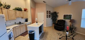 A kitchen with cabinets, a sink, a trash bin, a small island, and a dining area with a chair and table. - Right Direction Rentals 2 Bedroom Villa 9941 (Port St. Lucie)