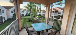 A balcony with a glass table and chairs, overlooking a courtyard with palm trees and apartment buildings. - Right Direction Rentals 2 Bedroom Villa 9941 (Port St. Lucie)