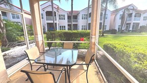 Un espace extérieur aménagé, abrité et doté d’une table et de chaises en verre, offrant une vue sur un jardin paysager et des immeubles résidentiels à plusieurs étages.