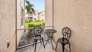 Un balcó amb dues cadires i una taula petita, amb vista a un edifici que té un jardí verd i un cel serè.
