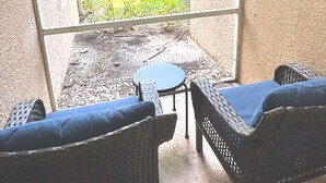 A balcony with two wicker chairs and a small table, overlooking a grassy area with trees. - Right Direction Rentals Studio Villa 9970 (Port St. Lucie)