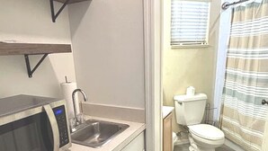 A compact kitchen with a microwave, sink, and dishwasher. A shower curtain is visible in the adjacent bathroom. - Right Direction Rentals Studio Villa 9970 (Port St. Lucie)
