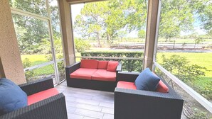 Een overdekt terras met rieten meubels en rode kussens, met uitzicht op een aangelegde tuin.