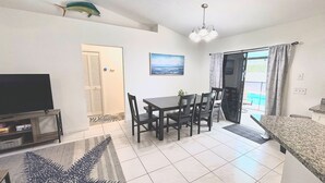 A living room with a TV, dining table, chairs, a painting, and a sliding glass door to a pool. - Right Direction Rentals Owl's Oasis (Port St Lucie)