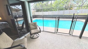 A room with a glass door leading to a swimming pool, a chair, and a bed. - Right Direction Rentals Owl's Oasis (Port St Lucie)