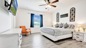 Une chambre à coucher comprenant un lit, une table de chevet, une chaise, une commode, un ventilateur de plafond, une fenêtre avec des rideaux et des éléments de décoration murale.