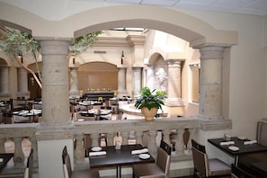 Un comedor interior con columnas de piedra, mesas y sillas, que presenta una fuente central y una planta en maceta.