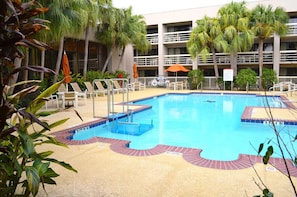 Una piscina con sillas de descanso, palmeras y un edificio de varios pisos al fondo.