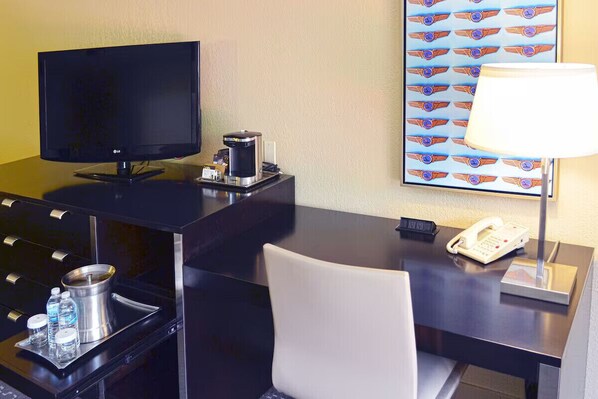 Ett hotellrum med ett skrivbord, en tv, en kaffebryggare, en telefon, en lampa och en tavla på väggen.