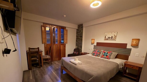 Une chambre à coucher avec un lit, un plancher en bois, une chaise, un bureau, une lampe et un téléviseur fixé au mur.