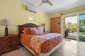 A bedroom with a bed, nightstands, a ceiling fan, an air conditioner, a wardrobe, and a sliding glass door leading to a balcony with a view of palm trees. - Ocean View Villa with Private Pool + Rooftop  and BBQ (Cabo San Lucas)