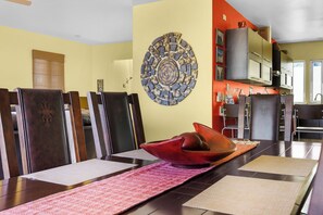 A dining area with a long table, chairs, a decorative wall piece, and a kitchen in the background. - Ocean View Villa with Private Pool + Rooftop  and BBQ (Cabo San Lucas)