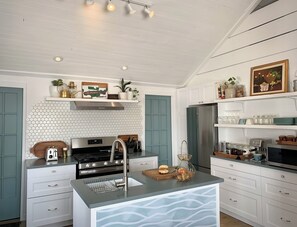 A kitchen with a central island, stainless steel appliances, and a tiled backsplash. - Saltbox by the Bay | Views, Bikes, Vetta Spa (Waubaushene)