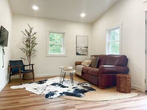 A living room with a brown leather sofa, a round coffee table, a small side table, a large window, and a wall-mounted TV. - The Duck House-Cozy 2 bedroom house in marvelous Crandon with AC (Crandon)