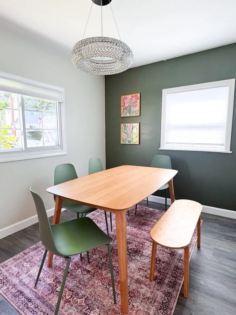 Un coin repas avec une table en bois, des chaises vertes, un banc, un tapis à motifs et des œuvres d’art accrochées aux murs.