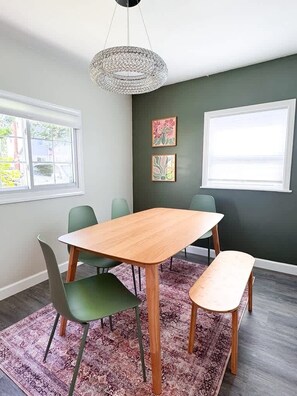 Et spiseområde med et træbord, grønne stole, en bænk, en gulvtæppe med et mønster og kunst på væggene.