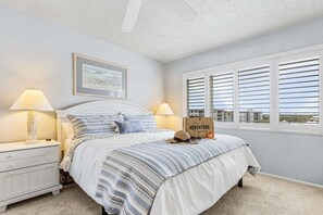 Une chambre à coucher avec un lit, deux tables de chevet, une lampe, un tableau encadré, une fenêtre avec des stores et un panneau sur la fenêtre.