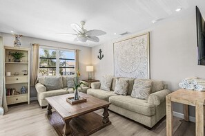 A living room with two sofas, a coffee table, a TV, a ceiling fan, and a large window. - Beachside Canal Retreat with Coastal Charm -  The Shore at Ocean Palms Retreat - Roelens (Fort Myers Beach)