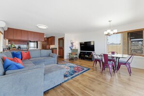 Ein modernes Wohnzimmer mit einem grauen Ecksofa, einem Esstisch mit Stühlen, einem Flachbildfernseher, Holzschränken und einer Küchenzeile.