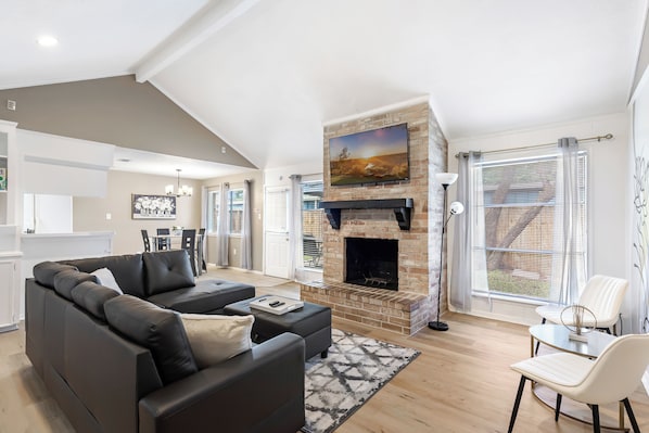 A modern living room with a fireplace, a sofa, a dining area, and a view of the outdoors.