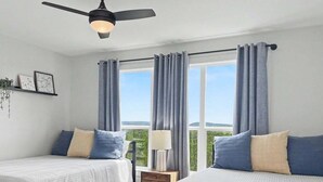 Una habitación con dos camas, un ventilador de techo, una ventana con cortinas, una mesita de noche y vistas a la vegetación.