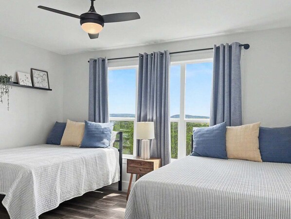 Quarto com duas camas, ventilador de teto, janela com cortinas, mesa de cabeceira e vista para o verde.