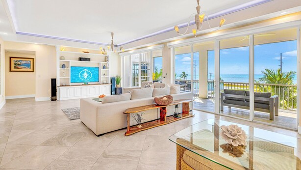 Een ruime woonkamer met een grote televisie, een bank, een glazen salontafel en een balkon met uitzicht op zee.
