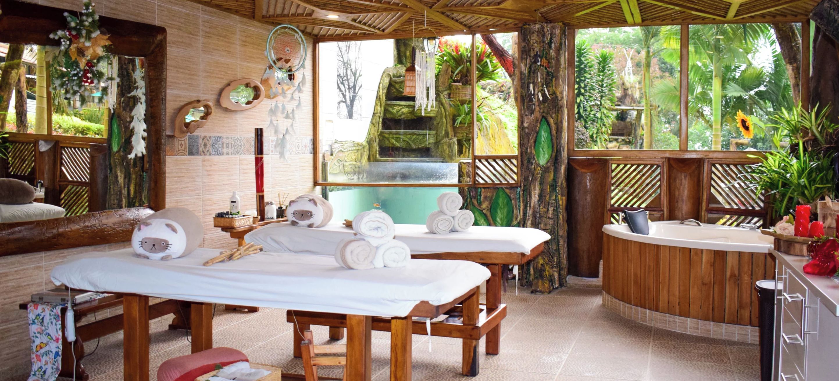 Ein Wellnessraum mit zwei Massagetischen, einem kleinen Waschbecken und einem Fenster mit Blick auf einen Garten.