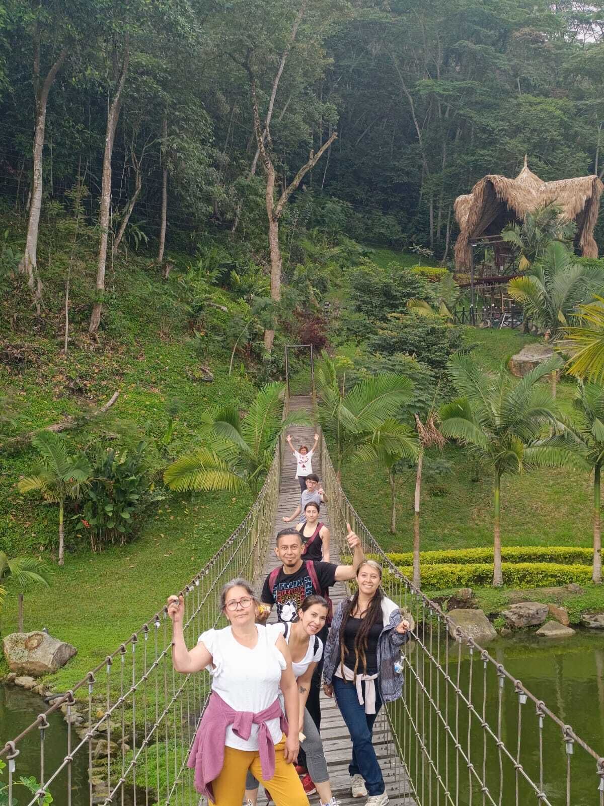 A group of people on a wooden suspension bridge over a river, surrounded by lush greenery and a thatched-roof structure.