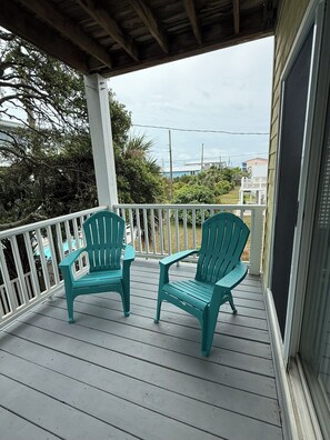 A covered porch with two turquoise Adirondack chairs, a white railing, and a view of the ocean and buildings in the background. - A'Boat Time-3 BR, 3 BA, beach retreat with backyard, 3 min walk to beach (Kure Beach)