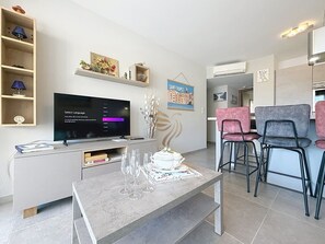Ein modernes Wohnzimmer mit einem Flachbildfernseher, einem Couchtisch mit Gläsern, einer dekorativen Vase und einem Essbereich mit Stühlen.