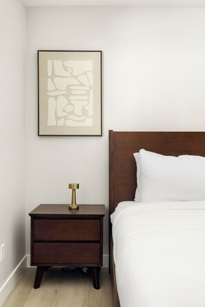 Un dormitorio con cabecera de madera, cama blanca, mesita de noche de madera con lámpara de latón, y una obra de arte abstracto enmarcada en la pared.
