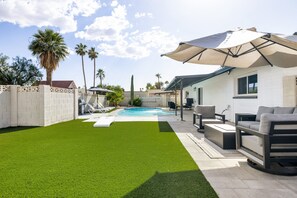 Un patio trasero con piscina, césped artificial, mobiliario de exterior y palmeras.
