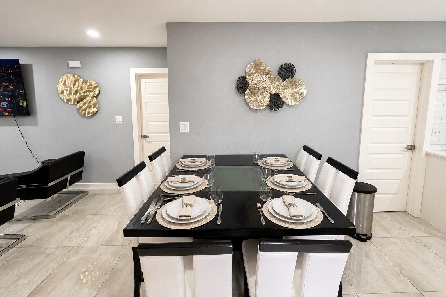 A dining area with a rectangular table set for six, white chairs, a black table, and a grey wall with decorative wall art.
