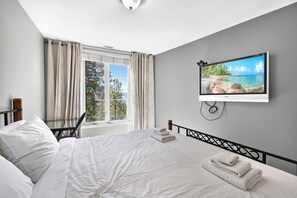 A bedroom with a bed, a desk, a chair, a TV, and a window with curtains. - Mountain View 3 Bd | Pool, Hot Tub & Golf 2208 (Kelowna)